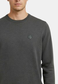 Maglione grigio scuro a maniche lunghe, con scollo rotondo e un piccolo logo circolare sul petto sinistro. Tessuto liscio, vestibilità casual.