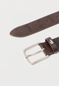 Καφέ δερμάτινη ζώνη με γεωμετρικό ανάγλυφο σχέδιο, ασημένια αγκράφα με χαραγμένο το όνομα της μάρκας και μεταλλικό λογότυπο κροκόδειλου στο βρόχο της ζώνης.