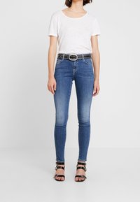 Jeans slim de mezclilla azul con un acabado desgastado, combinados con una camisa blanca de manga corta y un cinturón negro adornado, finalizados con tacones negros de tiras.