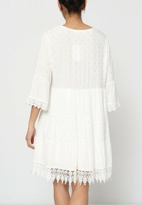 Robe blanche à volants avec des manches courtes en forme de cloche, ornée d'une bordure en dentelle au bas et de petits accents dorés dans le tissu. Design léger et fluide.