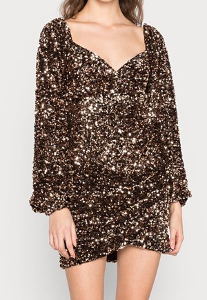 Kvinde iført en tætsiddende minikjole med lange ærmer, dækket af skinnende mørkebronzefarvede og gyldne pailletter, med en hjerteformet udskæring.