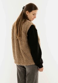 Beige nepbont vest met een gestructureerd oppervlak, oversized ontwerp en een contrasterend zwart langemouwen shirt eronder, tegen een neutrale achtergrond.