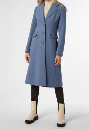 Femme portant un manteau bleu longueur genou avec trois boutons, poches avant à rabat, pantalon noir et bottines couleur crème.