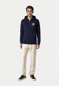 Felpa con zip di colore blu navy, con un logo bianco circolare sul petto, abbinata a pantaloni color crema e sneaker nere. Tessuto morbido, vestibilità rilassata.