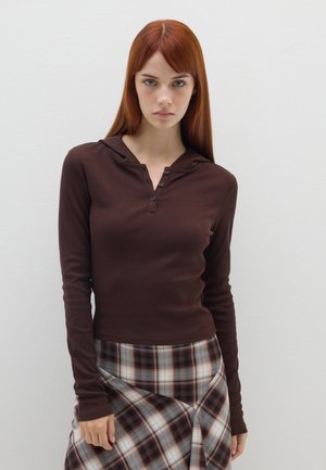 Bruine lange mouw top met een capuchon en knopdetails, gecombineerd met een geruite rok in tinten van rood, blauw en wit, op een lichte achtergrond.