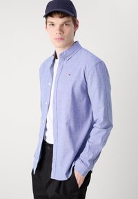 Camisa de botones azul claro hecha de una tela texturizada, con un pequeño logo en el pecho, llevada sobre una camiseta blanca y con pantalones negros.