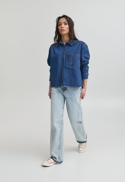 Mörkblå denimskjorta med en bröstficka, kombinerad med ljusblå vida jeans och vita sneakers, stående mot en neutral bakgrund.