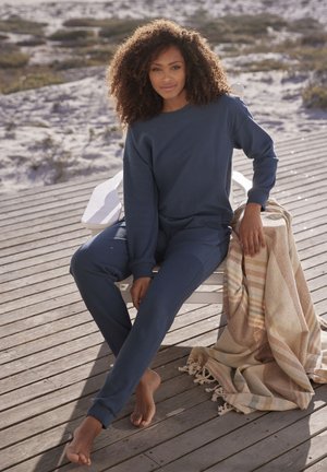 Marineblaues geripptes Loungewear-Set mit langen Ärmeln und Hosen, kombiniert mit einer gewebten Decke mit Streifen und Fransen, auf einer Holzterrasse.