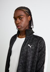Zwarte zip-up jas met witte accenten, voorzien van een logo en een overal patroon. Draaglijk over een strakke witte tanktop. Soepele textuur.