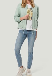 Woman wearing light green zip-up jacket, white graphic t-shirt, light blue skinny jeans, and beige sneakers against a plain background.