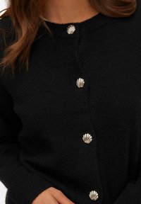 Svart stickad kofta med gyllene knappar formade som sjöskal, buren av en person med ljust brunt hår, handen vilar nära midjan.