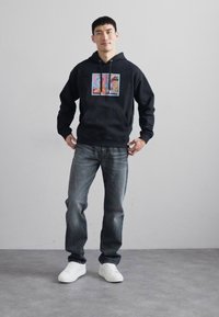 Black hoodie with a colorful printed graphic on the front, paired with faded blue jeans and white sneakers on a gray floor.