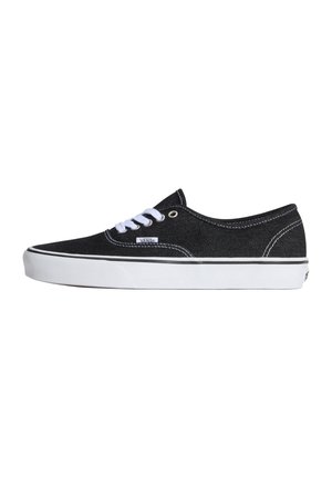 Deportiva Vans negra de corte bajo con suela blanca de goma, cordones blancos y etiqueta visible de la marca en el lateral, vista desde el exterior.