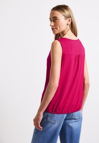 Fuchsiový top bez rukávů s kulatým výstřihem a nařaseným elastickým lemem, v kombinaci se světle modrými širokými džínami. Hladký povrch látky.