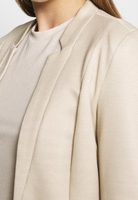 Beige rippad kavaj med en slät textur och strukturerad krage, bärs över en ljusbeige crewneck-tröja, vilket visar ett lager-på-lager-design.