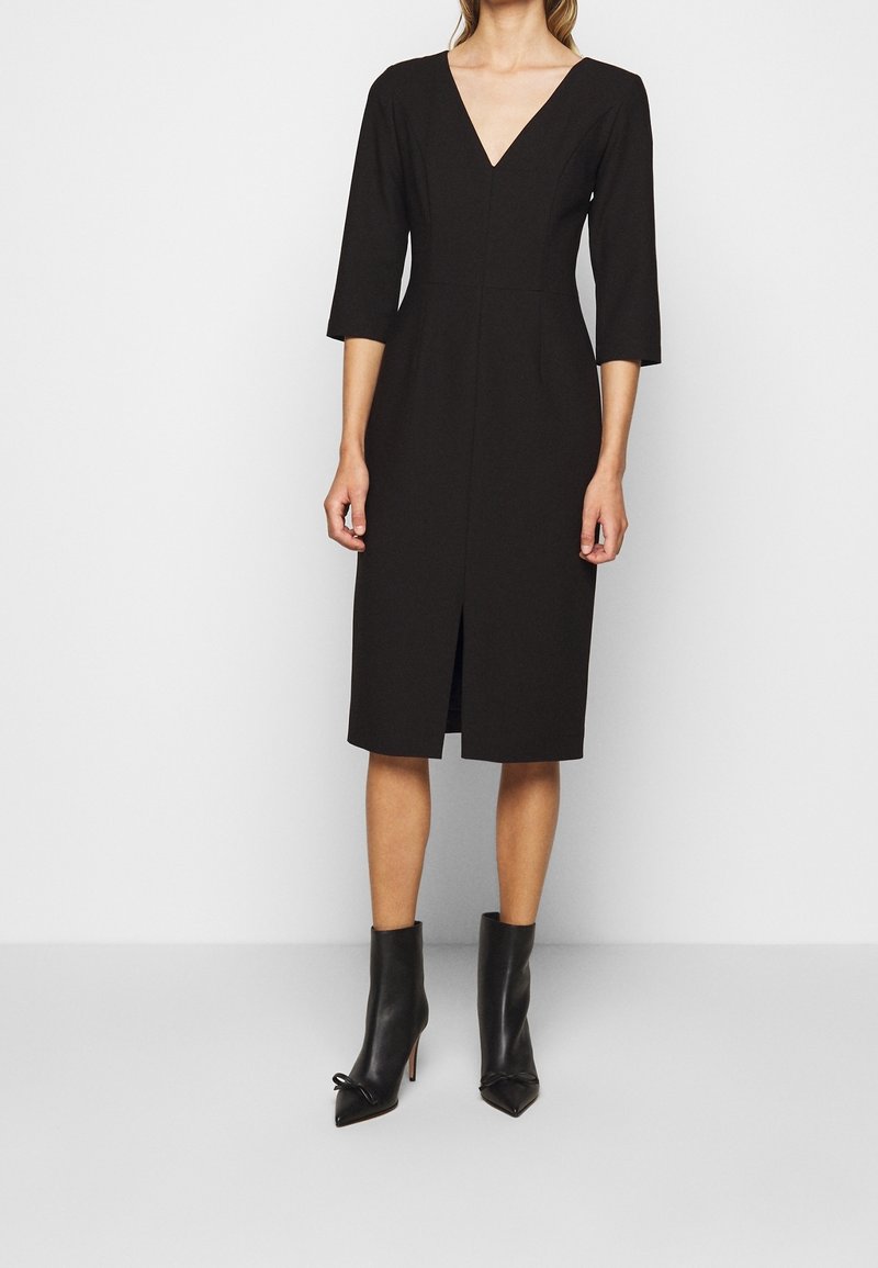 Femme portant une robe noire longueur genou avec manches trois quarts, décolleté en V, fente à l’avant et bottines noires à talons.