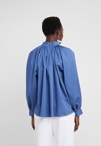 Femme portant un chemisier bleu ample avec un tissu froncé à l'arrière et un pantalon blanc, vue de dos sur un fond uni.