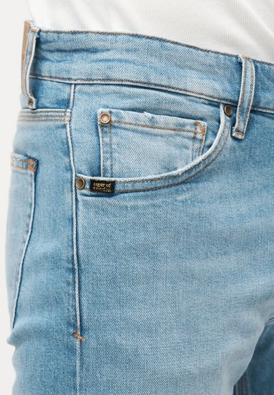 Φωτεινό μπλε τζιν με ορατά μεταλλικά κουμπώματα στην τσέπη και μια μικρή μαύρη ετικέτα ραμμένη κοντά στη ραφή της μπροστινής τσέπης.