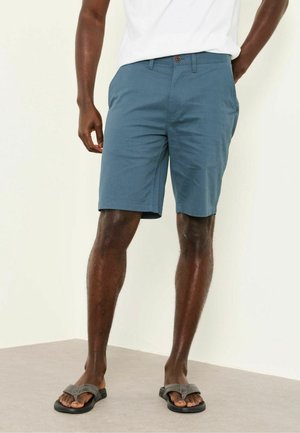 Mann trägt türkisfarbene kniehohe Shorts, weißes T-Shirt und graue Flip-Flops, steht vor einem schlichten hellen Wand- und Bodenhintergrund.