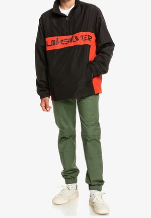 Sort og rød quarter-zip pullover med "Quiksilver"-logo, kombineret med grønne cargojoggers og hvide sneakers på en stående person.