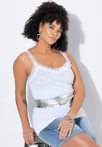Top bianco senza maniche con rifiniture in pizzo e decorazioni in strass, abbinato a una cintura metallica argentata su una camicia bianca a strati e pantaloni corti di jeans.