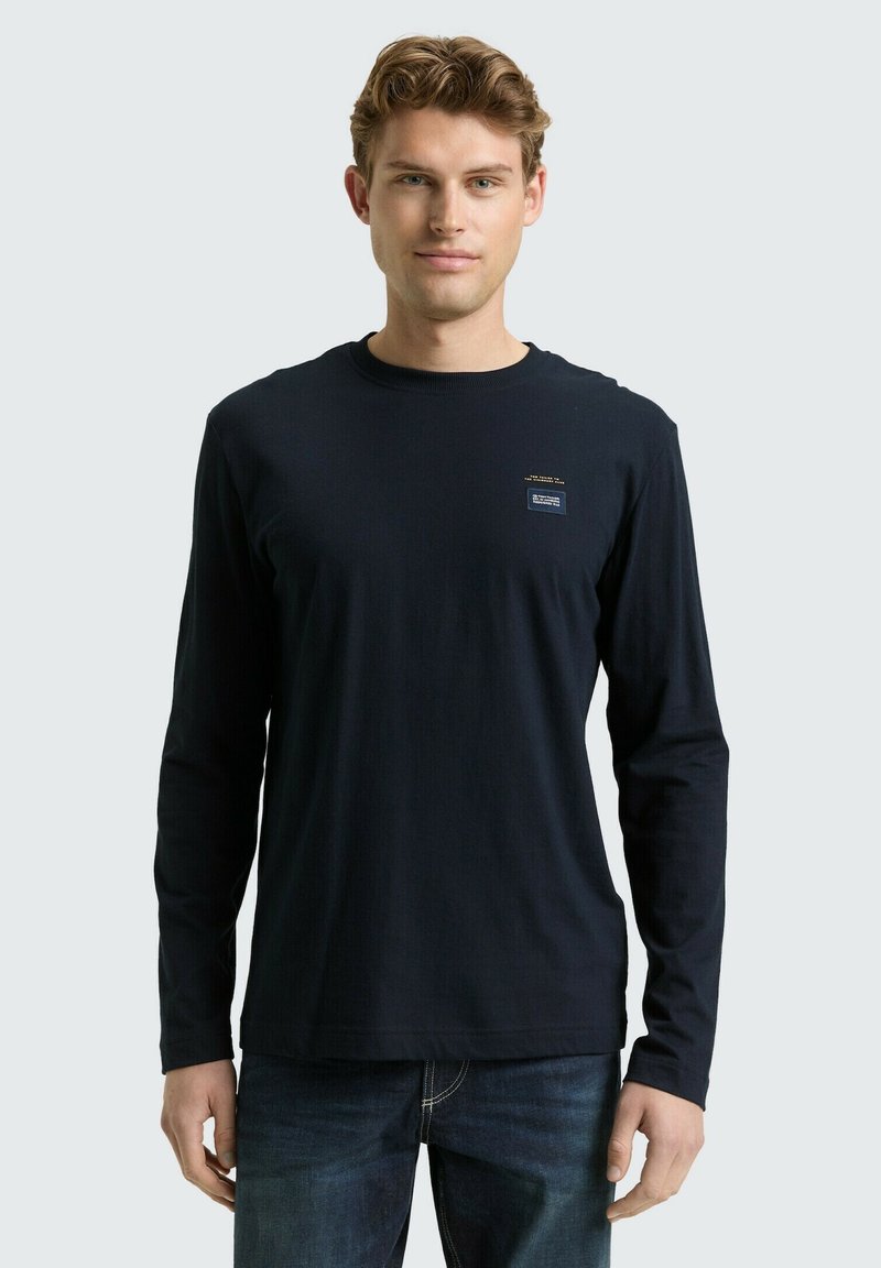 Granatowa koszula z długim rękawem wykonana z miękkiego materiału, z okrągłym dekoltem i małym graficznym akcentem na piersi. Noszona z jeansami.