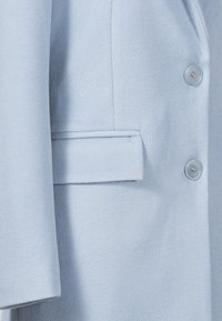Lichtblauwe op maat gemaakte blazer met een gestructureerde stof, notch-kraag, voorzak en knopen details. Zachte, gladde textuur is duidelijk zichtbaar.