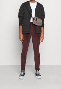Chaqueta negra acolchada, camiseta blanca, pantalones ajustados burdeos y zapatillas negras. Bolso cruzado en gris con cremalleras y correa.