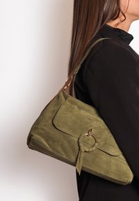 Una donna con un top nero porta una borsa a tracolla in suede verde oliva con un anello dorato e un dettaglio a nappina sulla tracolla.