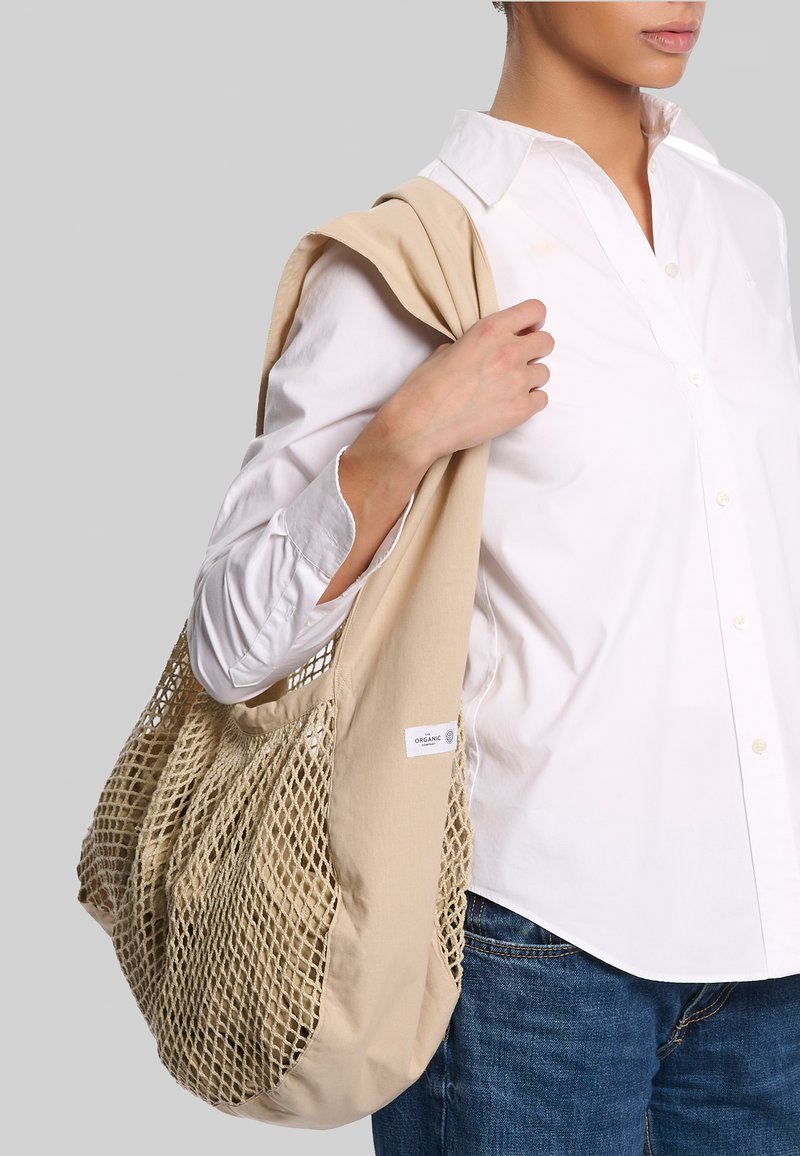 Person iført hvid skjorte og jeans bærer beige net-økologisk taske over skulderen mod grå baggrund.