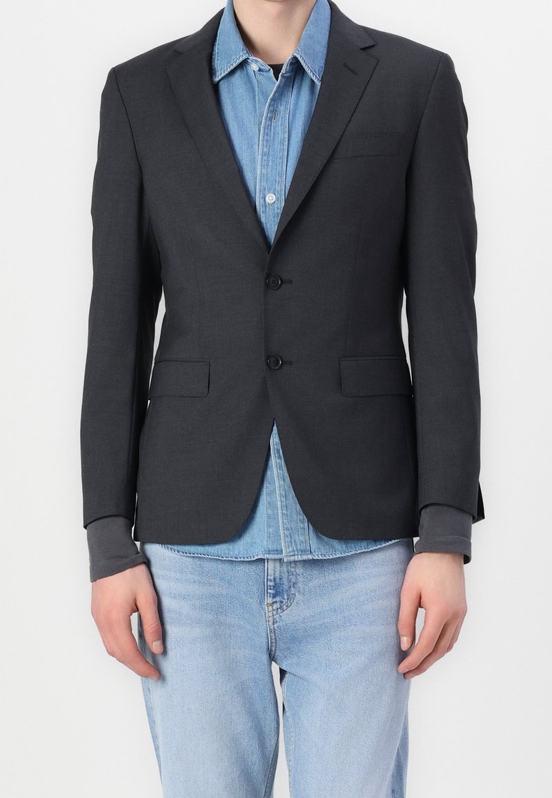 Dunkelgrauer Blazer aus strukturiertem Stoff mit zwei Knöpfen, aufgesetzten Revers und Fronttaschen, getragen über einem hellen Denimhemd.