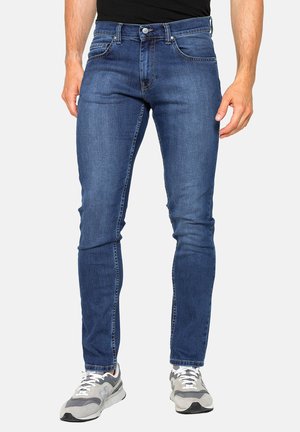 Uomo con jeans blu slim fit e scarpe da ginnastica grigie, in piedi davanti a uno sfondo chiaro e uniforme con le mani rilassate lungo i fianchi.