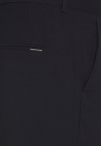 Nära vy av svarta, texturerade tygbyxor som visar en bakficka med en liten sydd etikett som säger "Scotch & Soda."