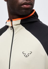 Κοντινό πλάνο ενός ατόμου που φοράει μπουφάν με φερμουάρ με μαύρα και μπεζ πάνελ και γιακά με πορτοκαλί περιθώριο, με λογότυπο προσώπου λεοπάρδαλης στο στήθος.