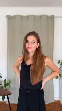 Mlada ženska z dolgimi rjavimi lasmi, oblečena v črno majico brez rokavov in hlače, stoji z rokami na bokih v svetli sobi s rastlinami in bež zaveso.