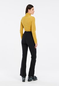 Top giallo a maniche lunghe abbinato a pantaloni neri attillati con aperture laterali. Il modello indossa stivali da sci scuri; sfondo minimalista.