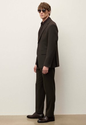 Moški v temno rjavi obleki, črni srajci in sončnih očalih, stoji ob bež steni z rokami sproščeno ob telesu.