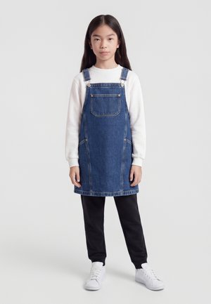 Jeune fille aux longs cheveux foncés portant un sweat-shirt blanc, une robe salopette en denim bleu, un pantalon noir et des baskets blanches, debout face à l’avant.