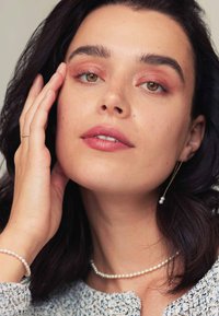 Collier et bracelet en perles avec de petites perles rondes. Gros plan sur un visage avec un maquillage léger et de longues boucles d'oreilles ornées de petites perles. Cheveux noirs.