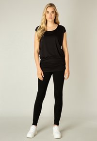 Zwarte top met korte mouwen en een relaxte fit, met een gathers in de taille, gecombineerd met zwarte legging en witte sneakers op een neutrale achtergrond.