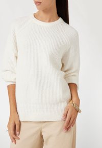 Pull ribbed blanc à manches trois-quarts et col rond. Présente un tricot texturé et un ourlet à motifs. Porté avec un pantalon beige clair.