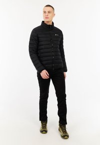 Jaunas vyras stovi, vilkėdamas juodą pūstą striukę, juodas kelnes ir žalias žygių batus, žiūrėdamas į šoną prieš baltą foną.