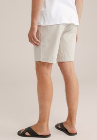 Beige pamut rövidnadrág egyenes szabással, lapos elülső résszel és felhajtott szegélyekkel; fekete papucsot viselve, hátulról nézve.