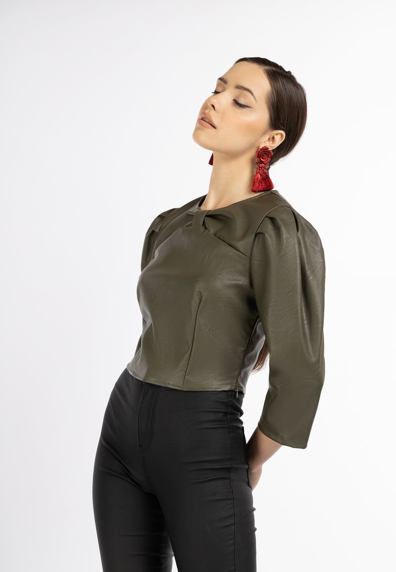 Olivegrüblouse aus Kunstleder mit Puffärmeln, einer Schleifendetail vorne, kombiniert mit schwarzen engen Hosen und roten Quastenohrringen.