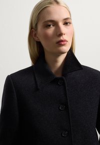 Manteau en laine noir avec un col haut, doté de deux grands boutons et d'une texture lisse. Le vêtement a un design minimaliste.