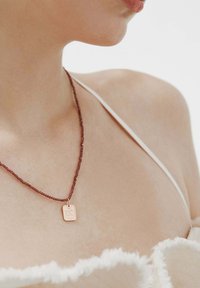Collar de bronce con un colgante rectangular, que presenta una superficie texturizada y una cadena de cuentas color burdeos, llevado sobre piel desnuda.