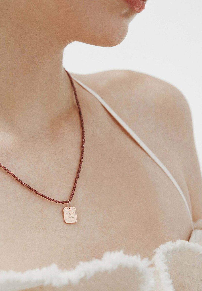 Collar de bronce con un colgante rectangular, que presenta una superficie texturizada y una cadena de cuentas color burdeos, llevado sobre piel desnuda.