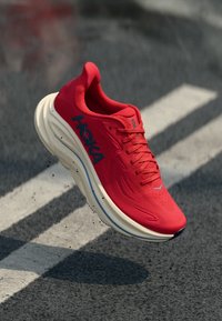 Κόκκινο αθλητικό παπούτσι με παχιά λευκή σόλα και μπλε λογότυπο "Hoka", κρεμασμένο στον αέρα πάνω από υφή ασφαλτοστρωμένου δρόμου με λευκές οδικές γραμμές.