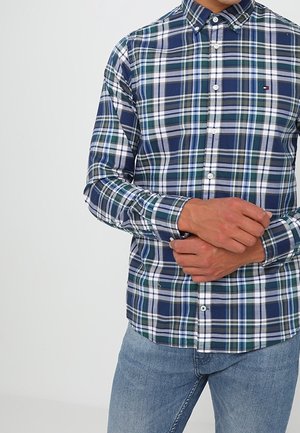 Muž v modré, zelené a bílé kostkované košili s knoflíkovým límečkem a světle modrých džínách, ruce sepnuté před sebou.