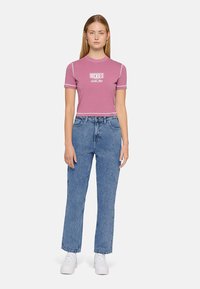 Jovem mulher em pé, vestindo uma blusa cropped rosa da Dickies, calças de ganga de cintura alta azuis e ténis brancos, contra um fundo claro e liso.